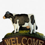 Black and white painted cast iron cow figurine with yellow grass base and brown welcome banner