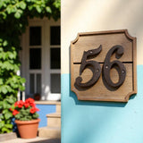 Cast iron house number 56 on wooden plaque with decorative rivets, mounted on blue cottage exterior