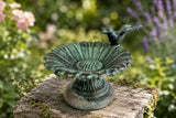 Verdigris cast iron sunflower bird bath with a perched bird on a weathered wooden stool in a flower garden