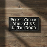 Black cast iron Please Check Your Guns at the Door saloon sign mounted on rustic wood