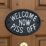 Cast iron oval plaque with white text Welcome Now Piss Off on dark finish mounted on door