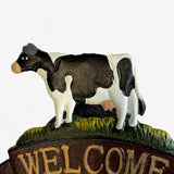 Black and white painted cast iron cow figurine with yellow grass base and brown welcome banner