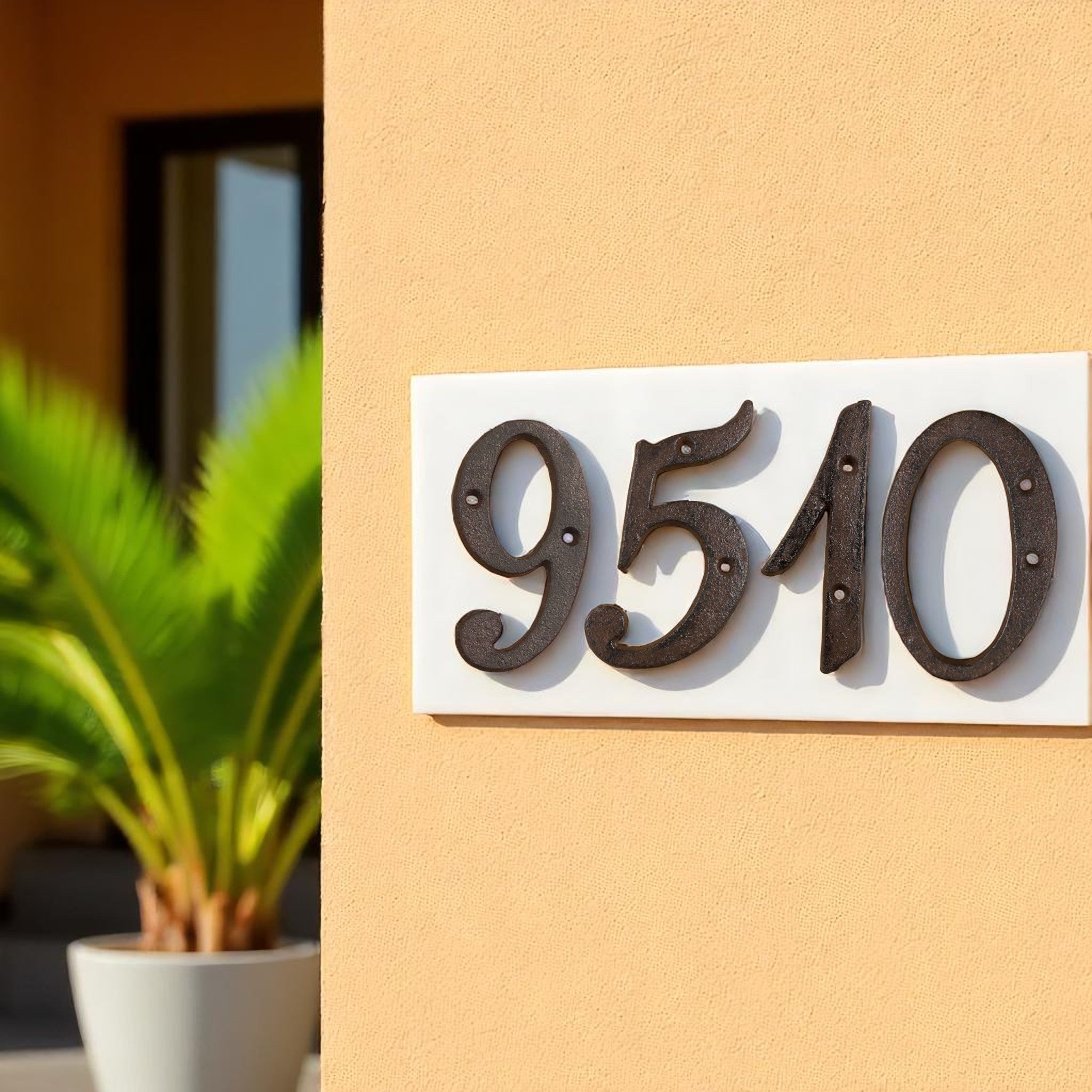 Antique cast iron house numbers 9510 on white wooden base, mounted on yellow stucco wall