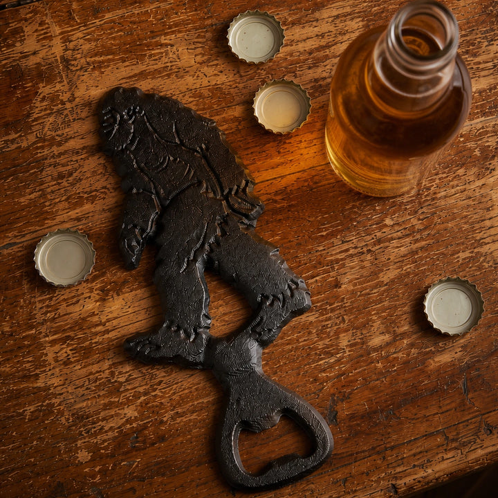 Black cast iron Bigfoot bottle opener on a wood bar with bottle caps, SKU GB78071